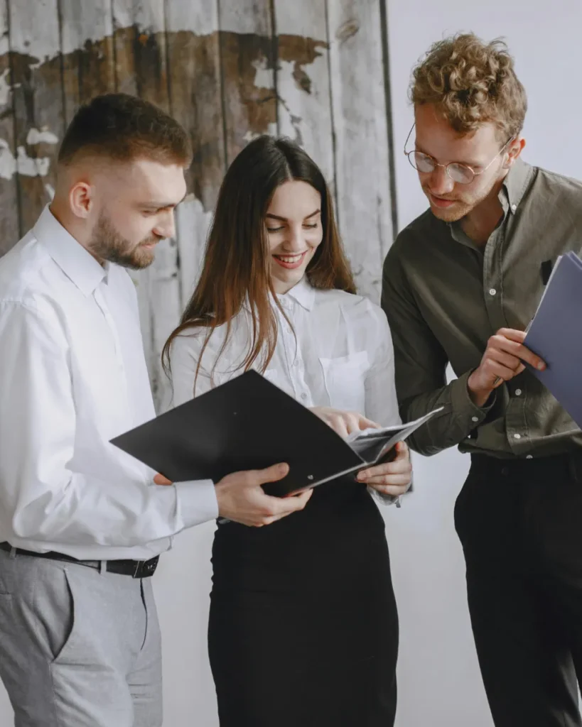 Das Bild zeigt 3 junge Businness-People die gemeinsam zusammen stehen, in eine geöffnete Mappe schauen und sich freundlich unterhalten und dient als Beitragsbild zum Thema "Jetzt bewerben- Jobangebote bei RGP Die Werbemittelprofis aus Berlin"